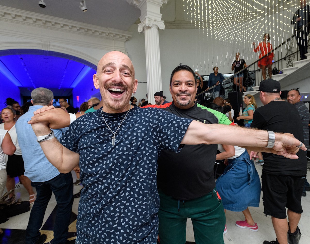 Two men smile and dance together in a room full of people.